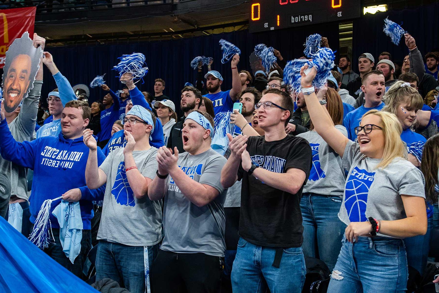 A large group of Sycamore fans, men and women, cheer for their team during a basketball game at the Hulman Center. They are clapping, cheering, holding blue and white mini-pompoms, and many are wearing light blue headbands. 